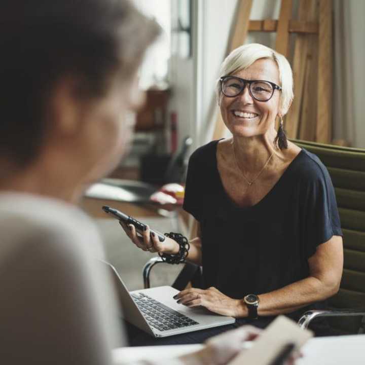 Middle aged woman working on phone and laptop with a colleague in an office