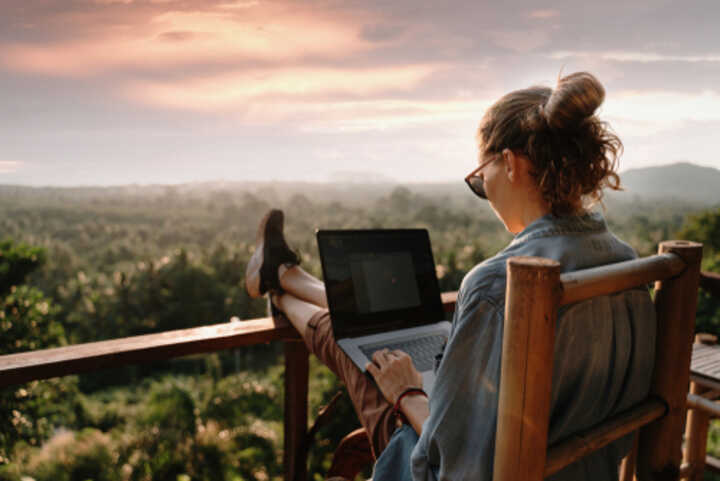 Young woman sitting on a balcony, working on a laptop, with a view of trees and a mountain