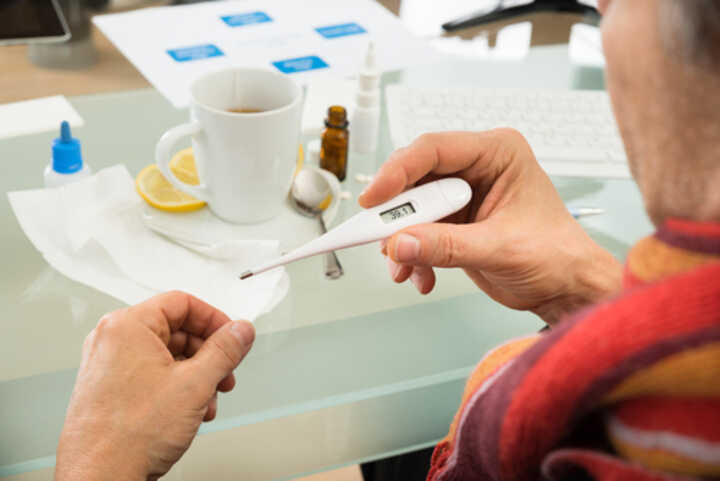 Sick businessman looking at a thermometer, with a hot drink on his desk